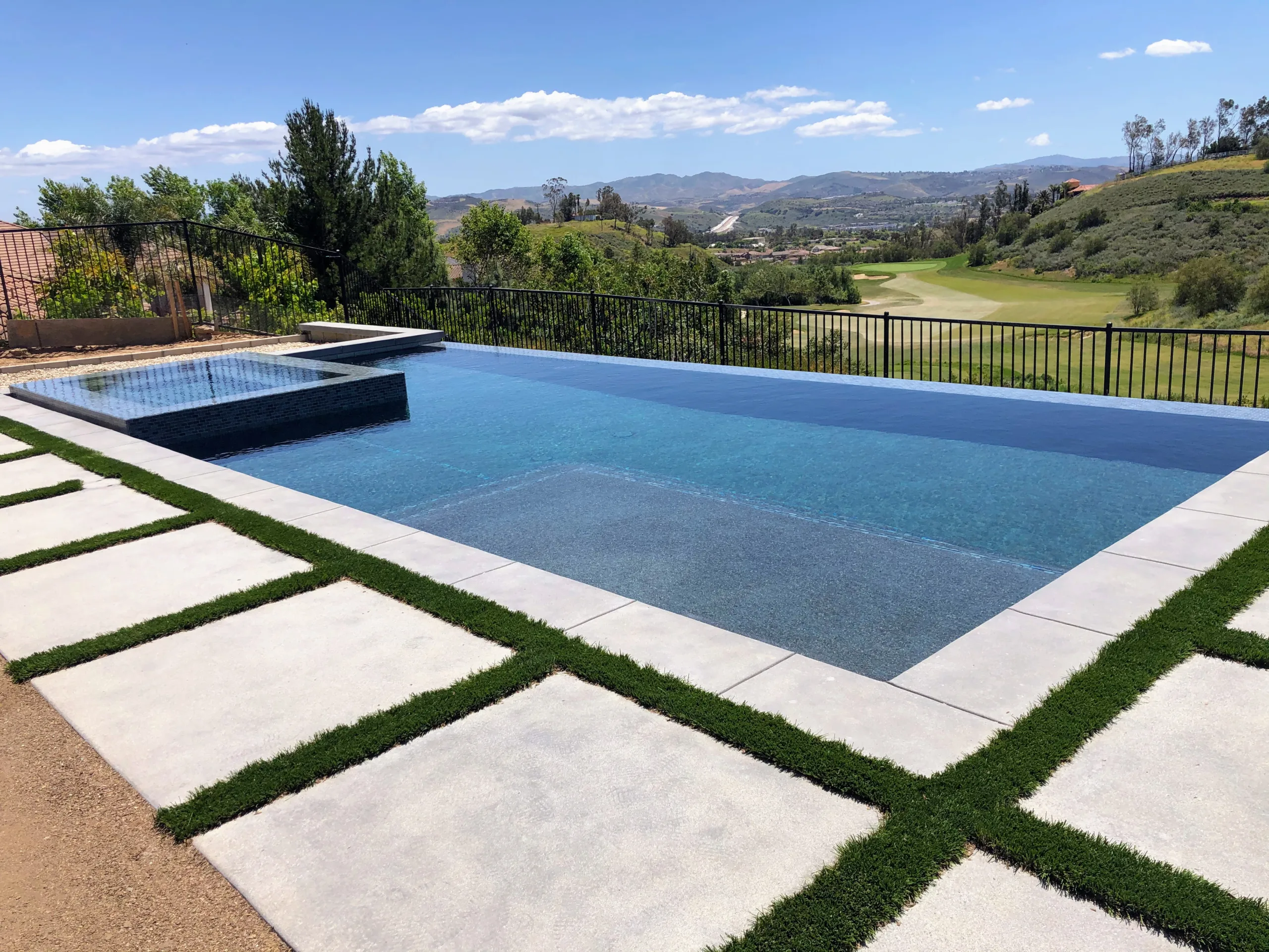 An infinity pool with adjacent hot tub overlooking a hilly landscape on a sunny day.