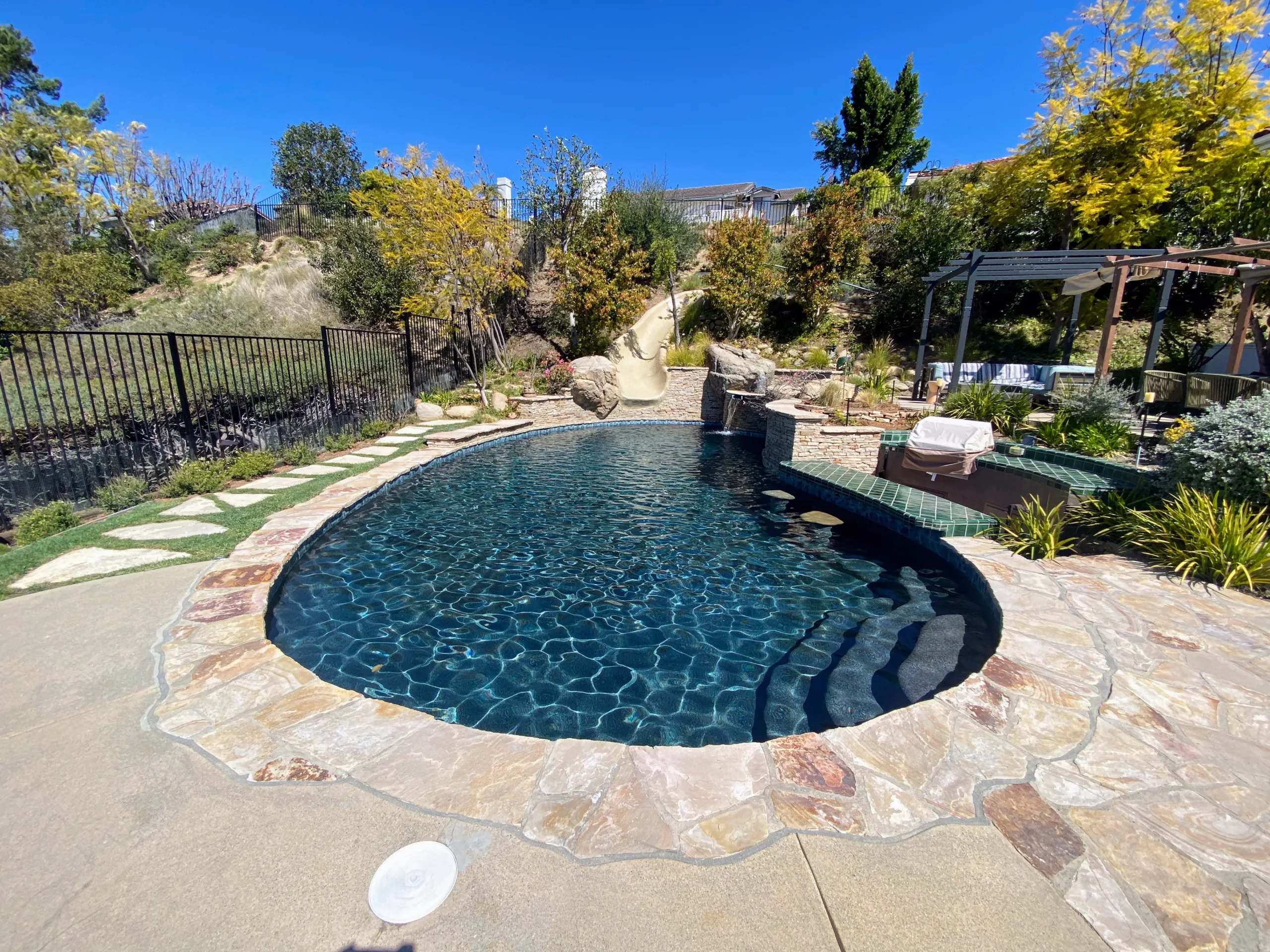 A backyard with a round swimming pool, stone edging, and surrounding plants. In the background, there's a slide, a pergola-covered seating area, and a fence. The sky is clear and blue.