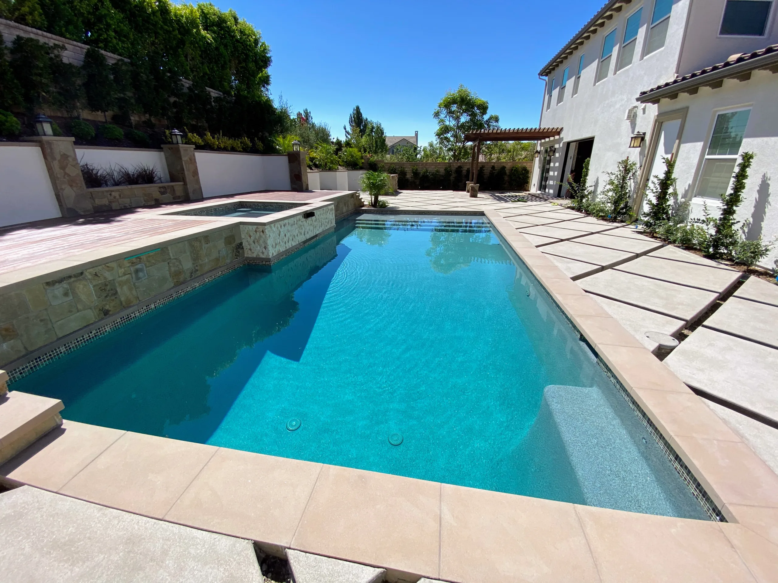 A modern backyard with a blue-tiled swimming pool, stone-edged hot tub, and paved patio surrounded by a white two-story house and greenery.